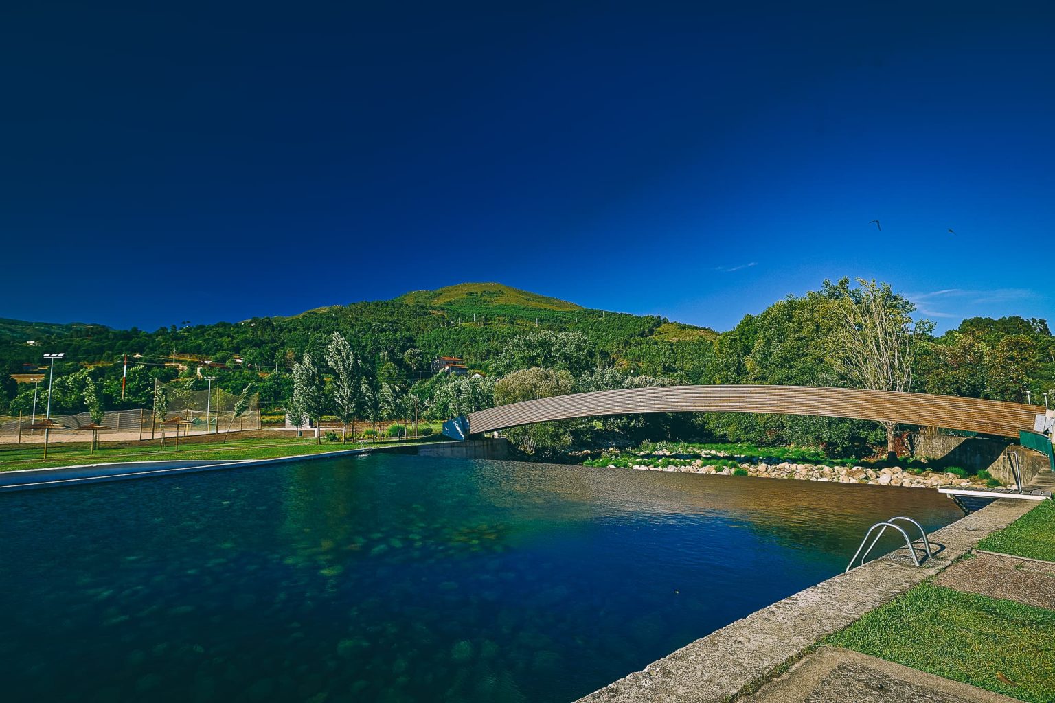 Praia fluvial com águas límpidas e uma piscina natural estruturada em Ribeira de Pena. No centro, uma moderna ponte pedonal de madeira em arco atravessa o rio, ladeada por margens relvadas, árvores e uma imponente montanha verde sob um céu azul limpo.